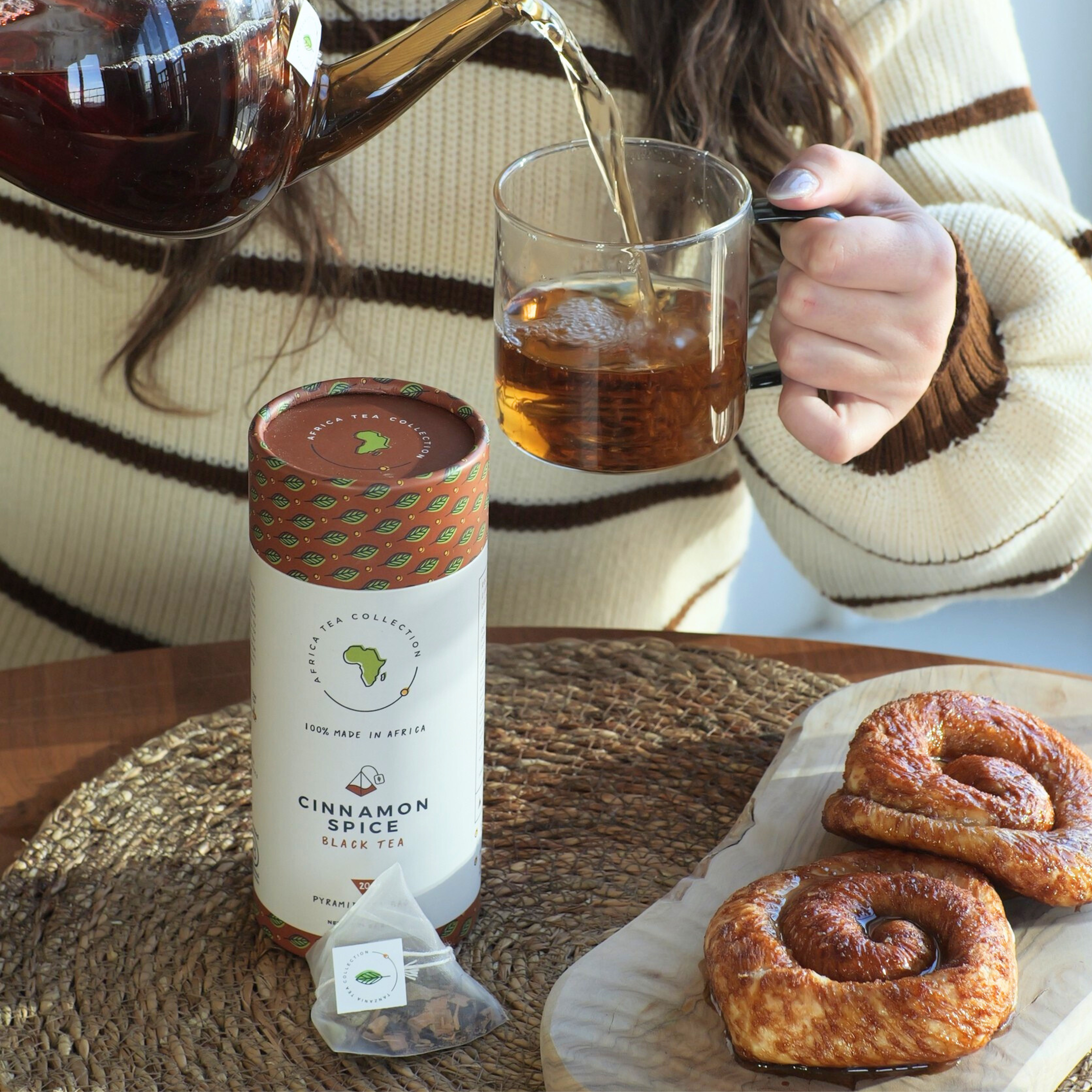 Person pouring tea into a glass mug with a cinnamon spice tea package and pastries on a table.