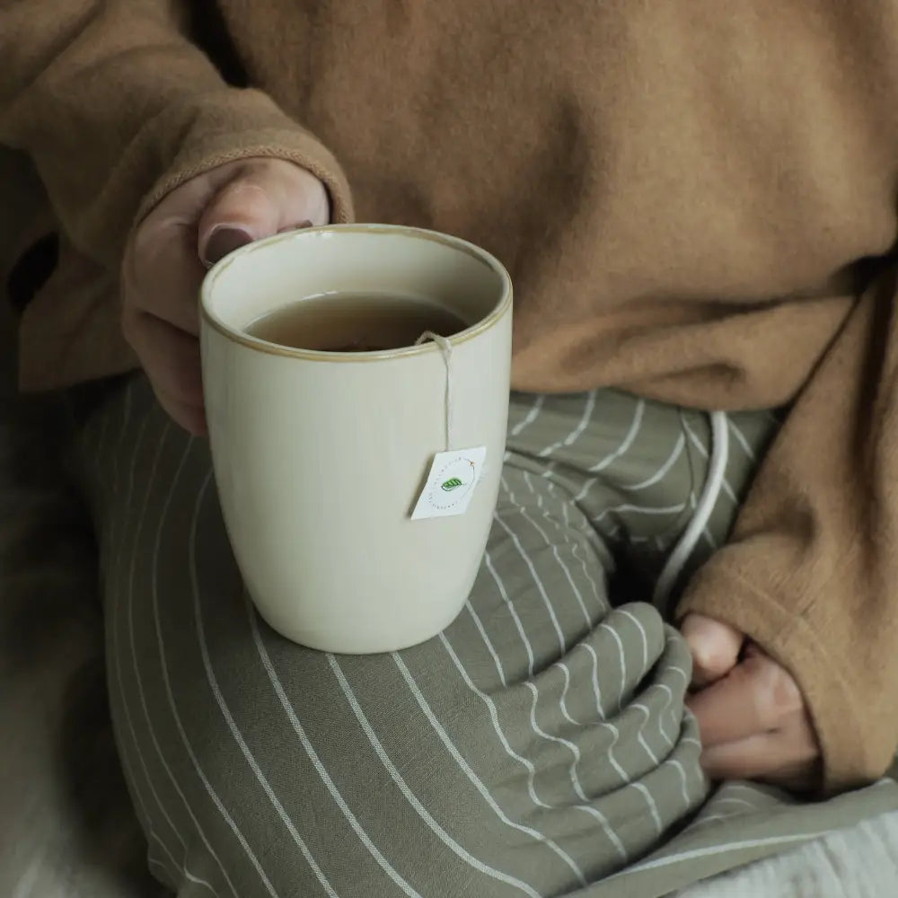 Person holding a white mug with a green leaf label on a striped cushion.