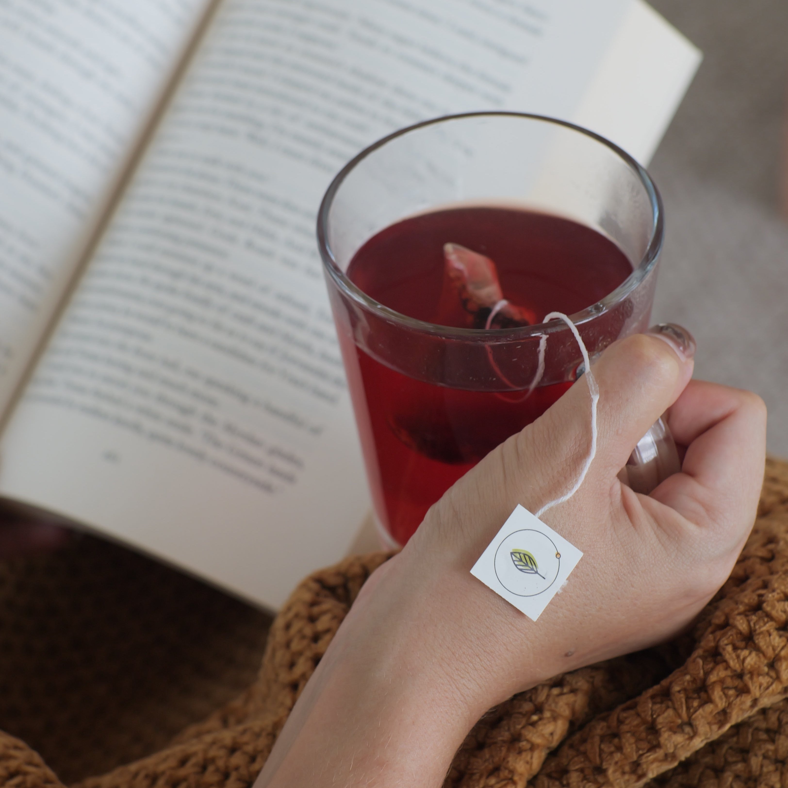 Hand holding a glass of tea with a teabag, next to an open book.