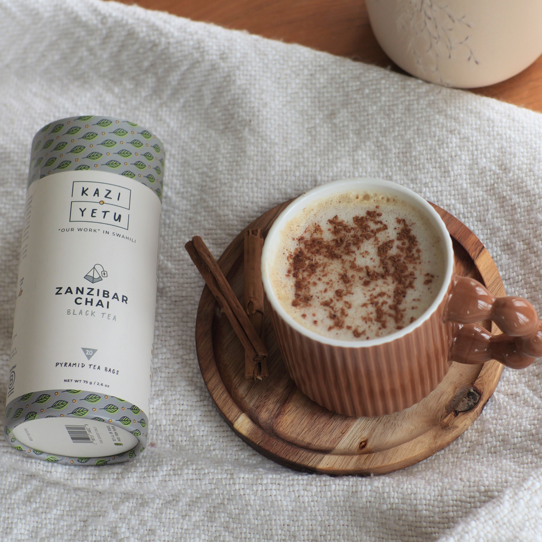 Kazi Yetu Zanzibar Zanzibar Chai tea canister with a cup of chai on a wooden coaster.