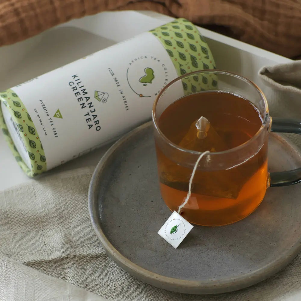 Glass of green tea with a tea bag on a ceramic plate next to Kilimanjaro Green Tea packaging.