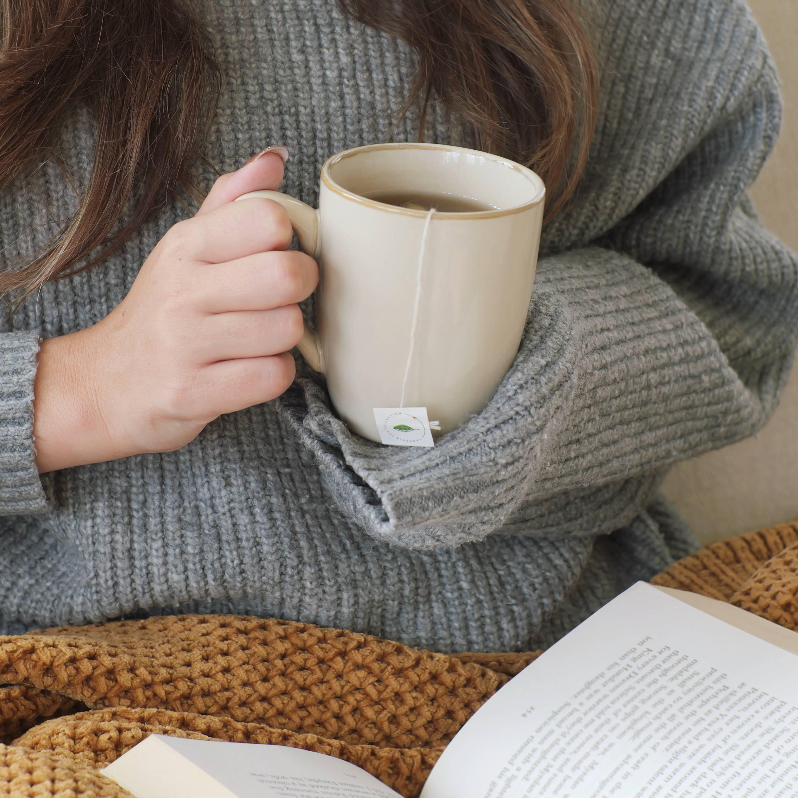 Person holding a mug with a tea bag, sitting on a brown textured surface with an open book.