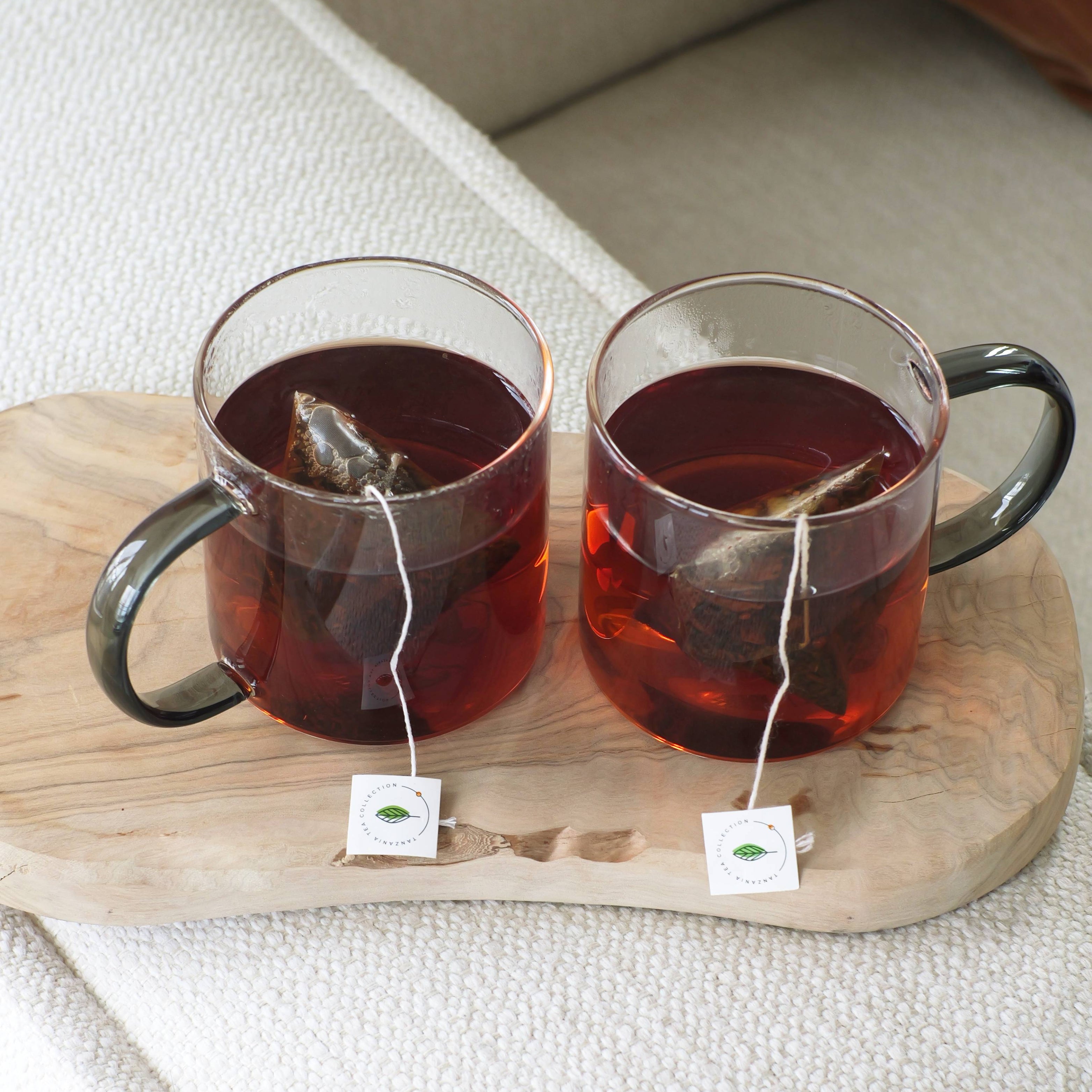 Two glasses of black tea with tea bags on a wooden board.