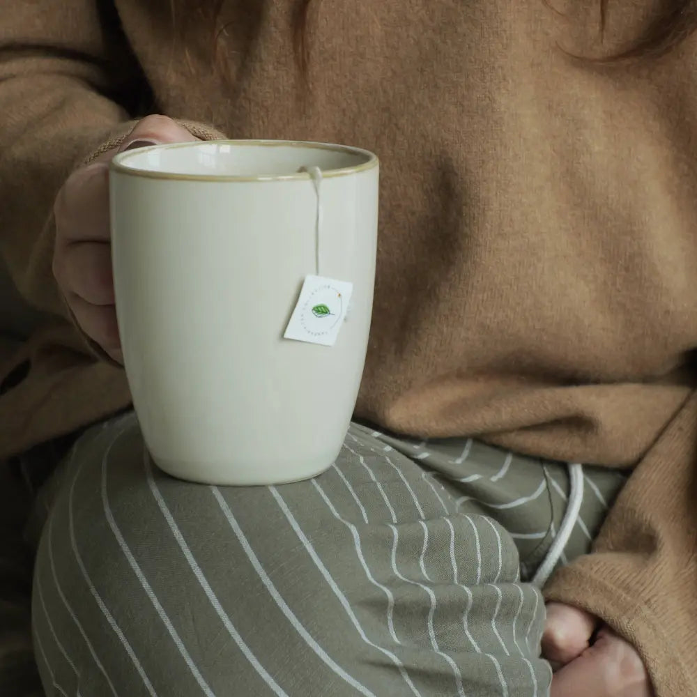 Person holding a beige mug with a tea bag tag, wearing a brown sweater and striped pants.